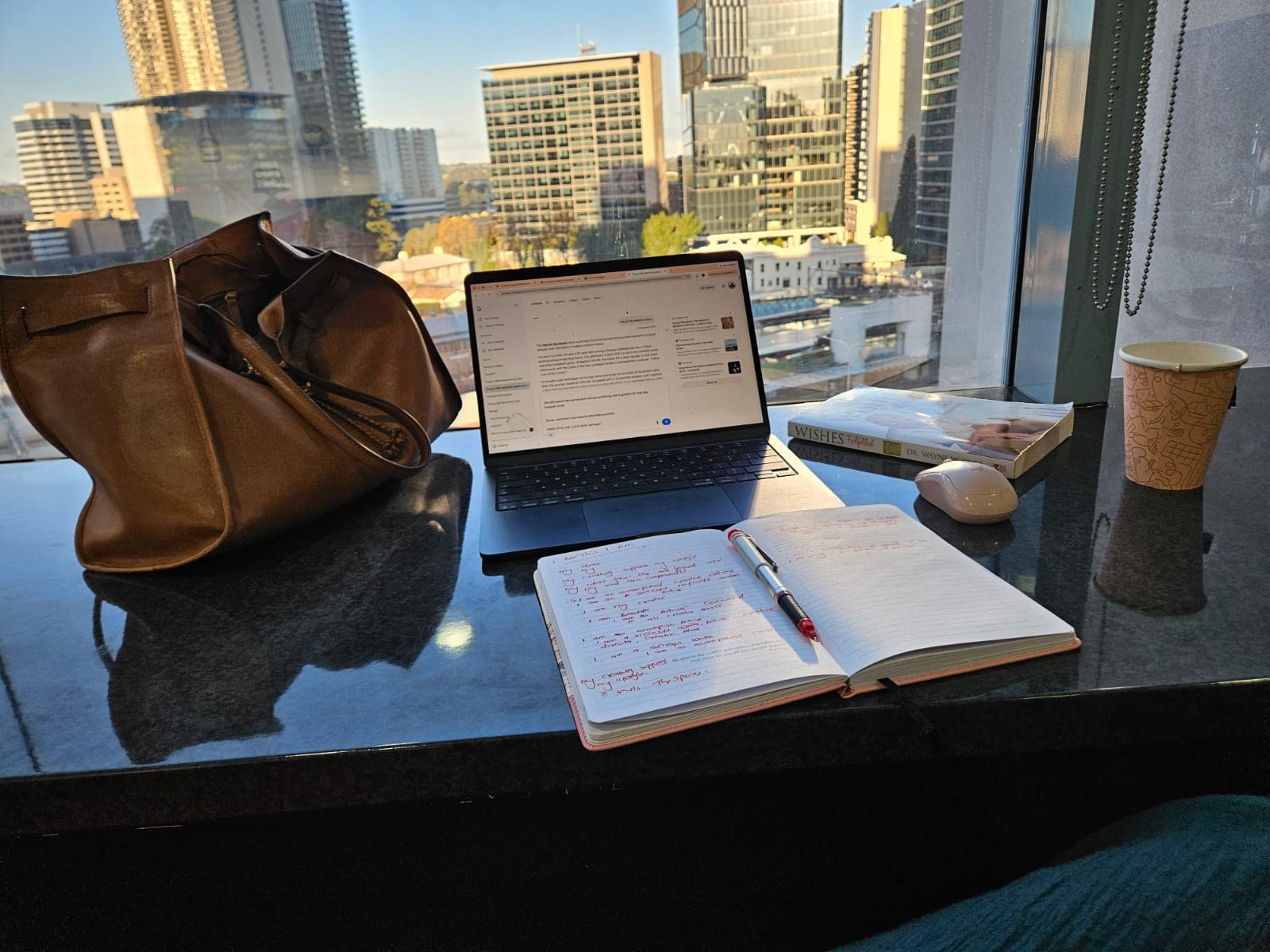 Rodah’s handwritten notebook, coffee, book and laptop on a dark reflective table