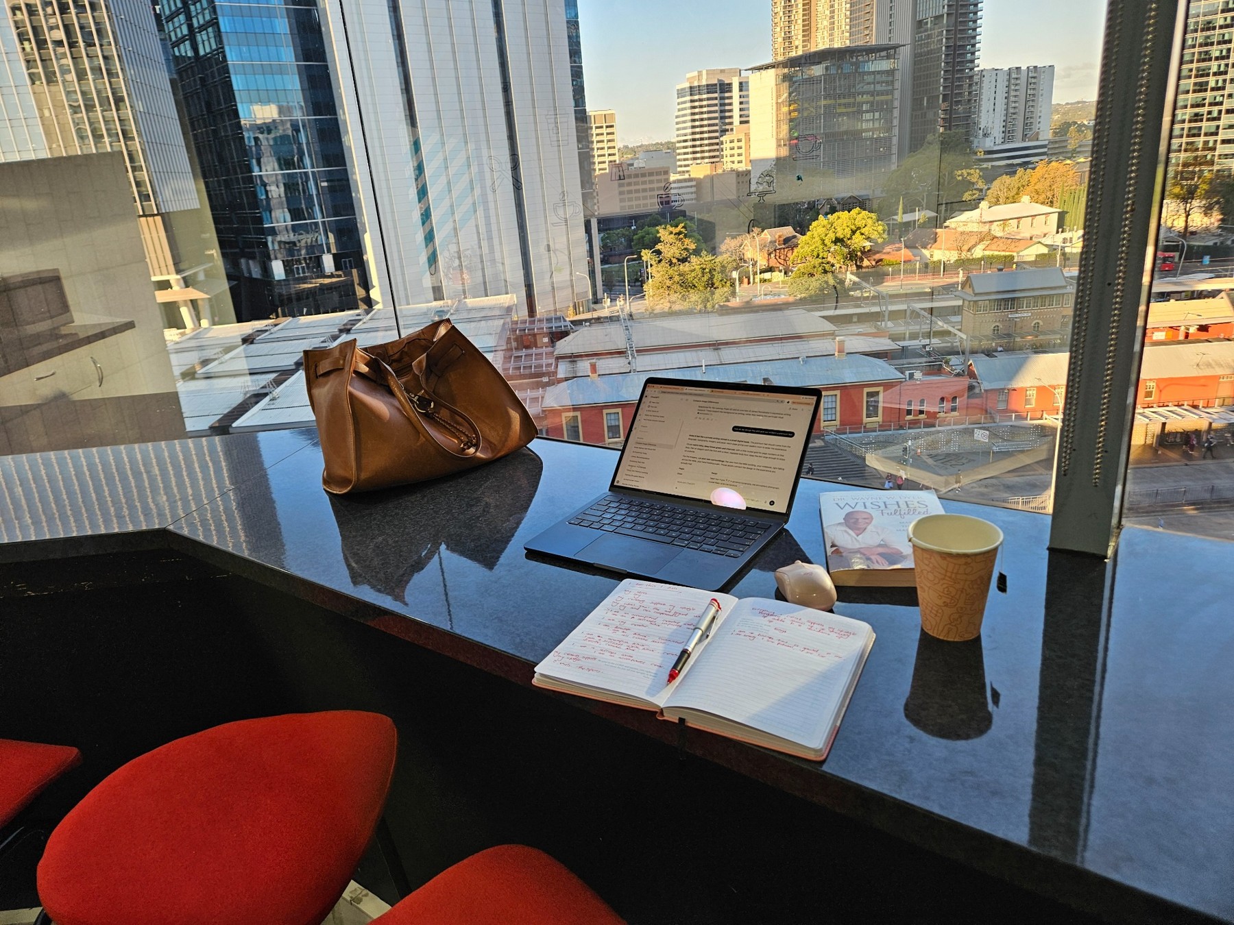 My notebook, coffee and laptop beside a city-facing library window